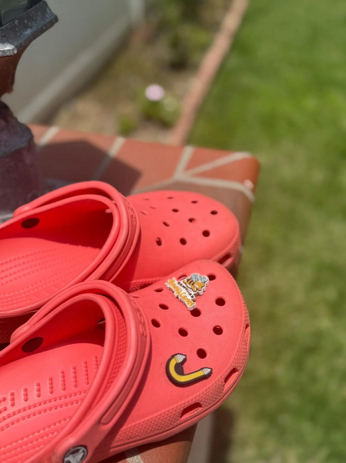 A pair of coral red Croc-style clogs with small decorative charms, photographed outdoors on a tiled surface with grass in the background.